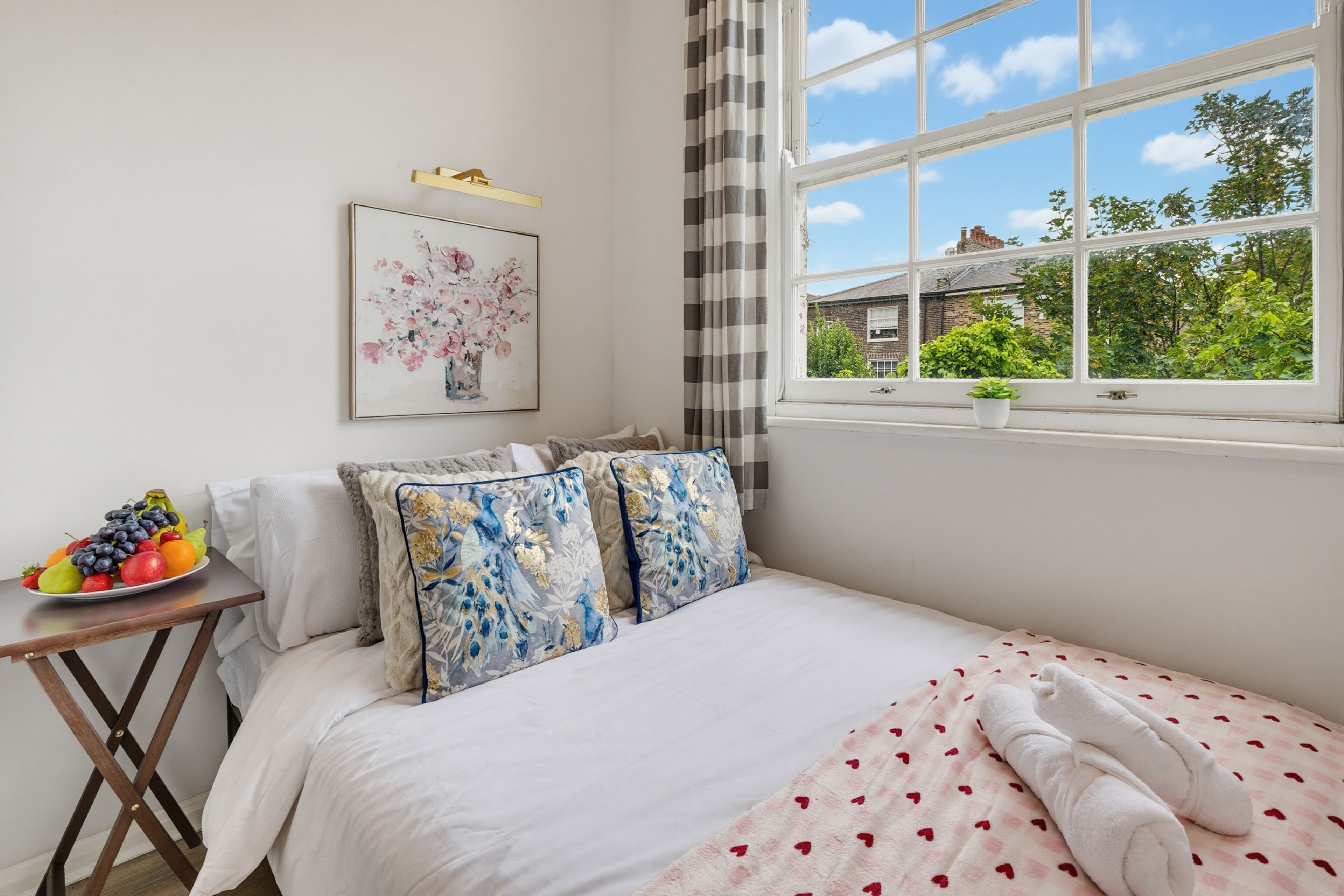 A bedroom with a bed, a bedside table with fruit, a window with a view of greenery, and a painting on the wall.