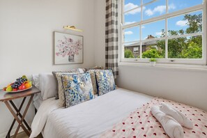 A bedroom with a bed, a bedside table with fruit, a window with a view of greenery, and a painting on the wall. - Arsenal Corner Studio, 4 minutes to Kings Cross, with Ensuite & Self Check-In (London)