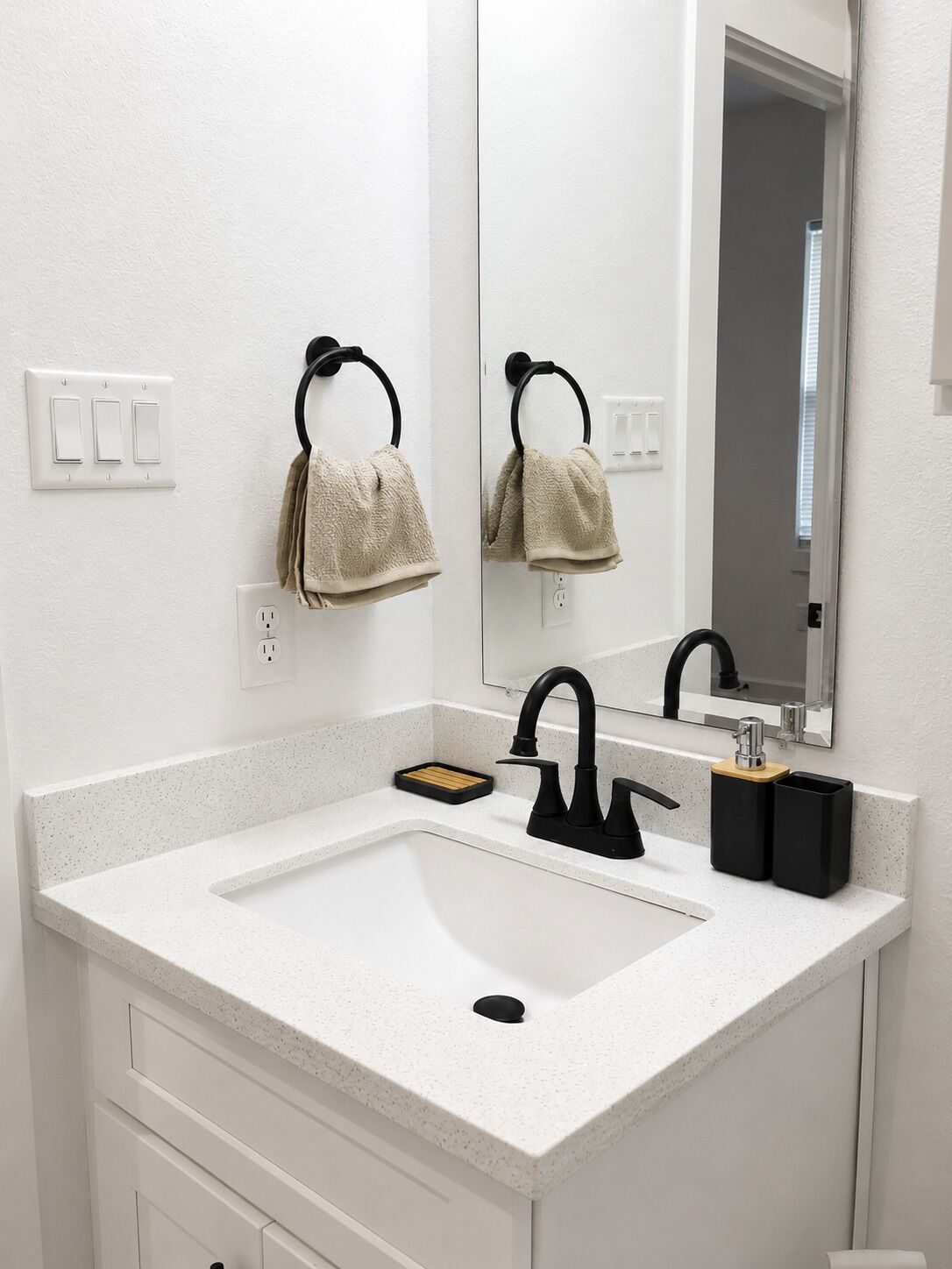 A bathroom with a white countertop sink, black faucet, and a mirror above. There are two towel rings on the wall, and a small wooden tray on the sink.