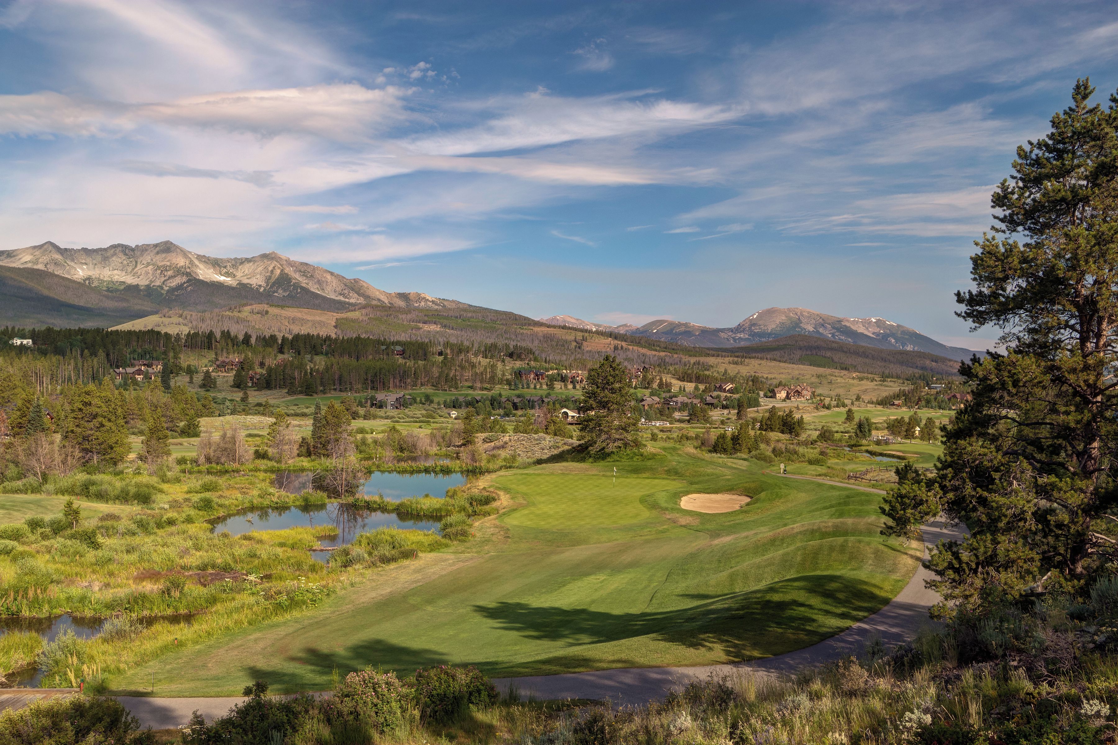 A golf course with a pond, surrounded by mountains and trees.