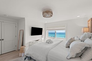 A bedroom with a bed, a television, a window with a view of the sea, and a ceiling light fixture. - 2BR Beachfront 30A Condo | Private Beach, Pool & Tennis (Seacrest)