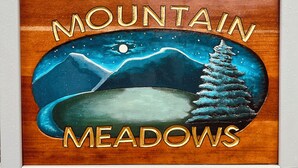 A framed artwork with the text 'MOUNTAIN MEADOWS' and a depiction of mountains, a tree, and a moon. - Warm and inviting condo 5 mins to Mt Snow-Dog friendly, 2 fireplaces, Great wifi (Wilmington)