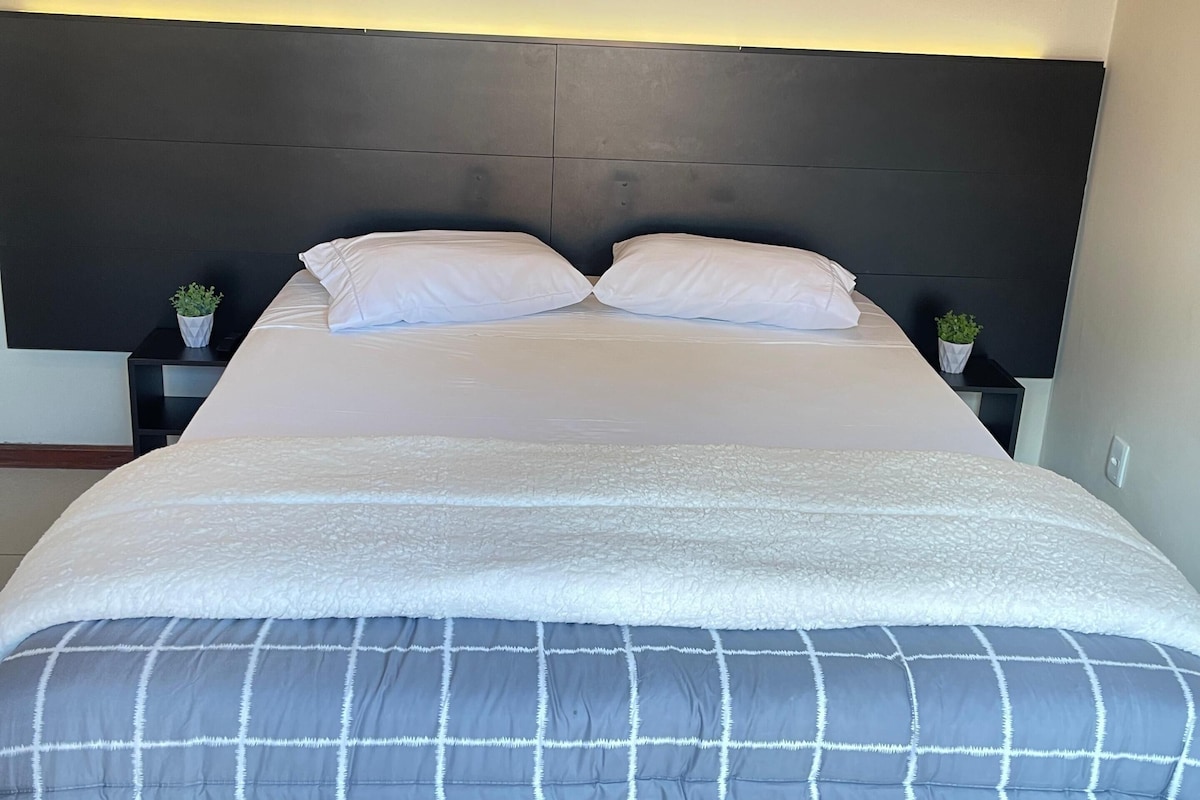 Een bed met een donker hoofdbord, witte kussens en een grijs-wit geruit laken.