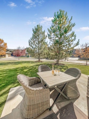 Un patio al aire libre con mobiliario de mimbre y una mesa, rodeado de un césped cuidado y árboles.