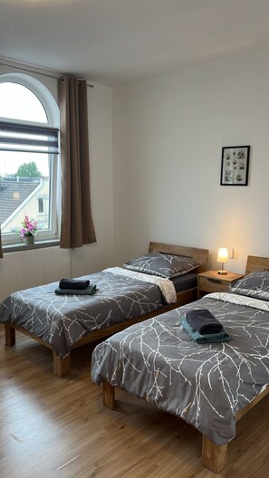 A bedroom with two beds, a window with curtains, a bedside lamp, and a framed picture on the wall. - Blunest Apartment 4: 4-Bedroom Apartment with Parking Space and Balcony (Bremerhaven)