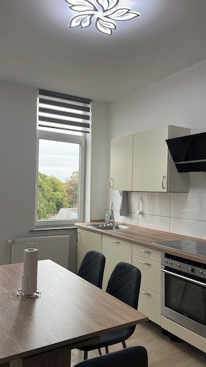 A modern kitchen with a dining table, chairs, a built-in oven, and a window with blinds. - Blunest Apartment 4: 4-Bedroom Apartment with Parking Space and Balcony (Bremerhaven)