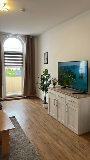A modern living room with a flat-screen TV, wooden cabinets, a potted plant, and a view of a coastal landscape through a large window. - Blunest Apartment 4: 4-Bedroom Apartment with Parking Space and Balcony (Bremerhaven)