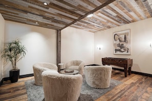 A room with a wooden ceiling, a round glass table, fluffy chairs, a small side table, a plant, and a painting on the wall.