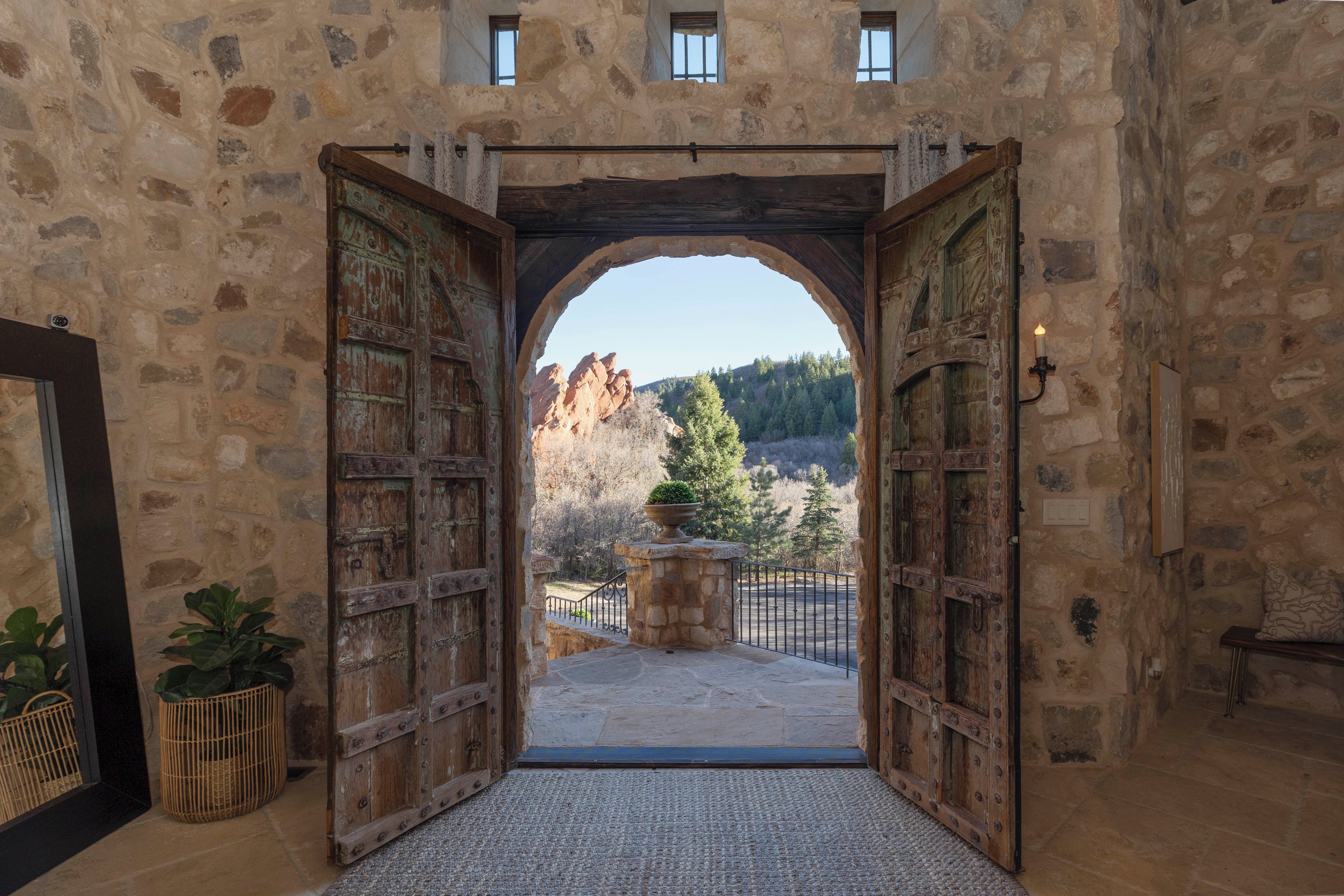 A stone building entrance with two large wooden doors, a stone wall, and a view of a mountainous landscape.