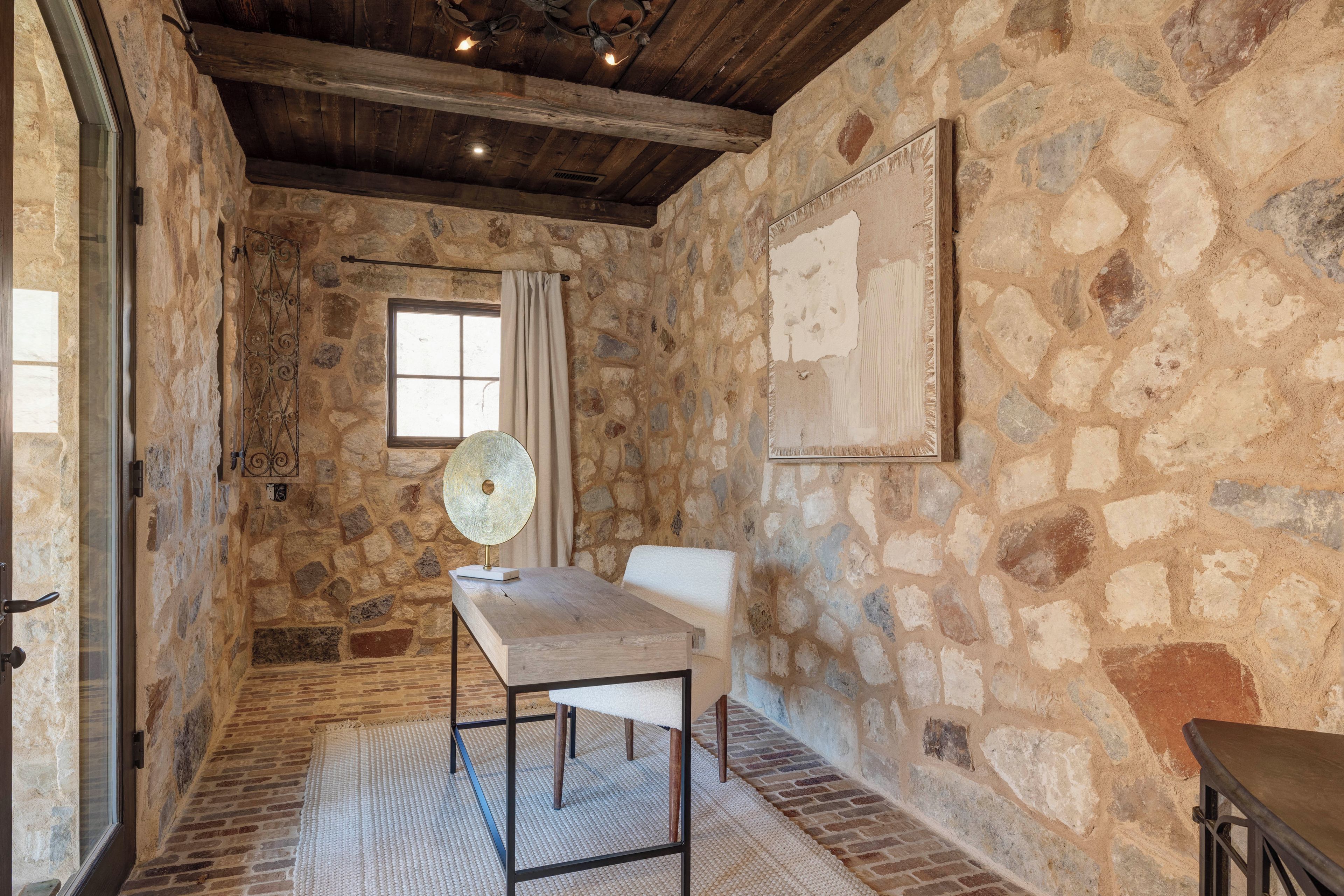 A stone-walled room with a wooden ceiling, a window with curtains, a table with a lamp, and a chair.