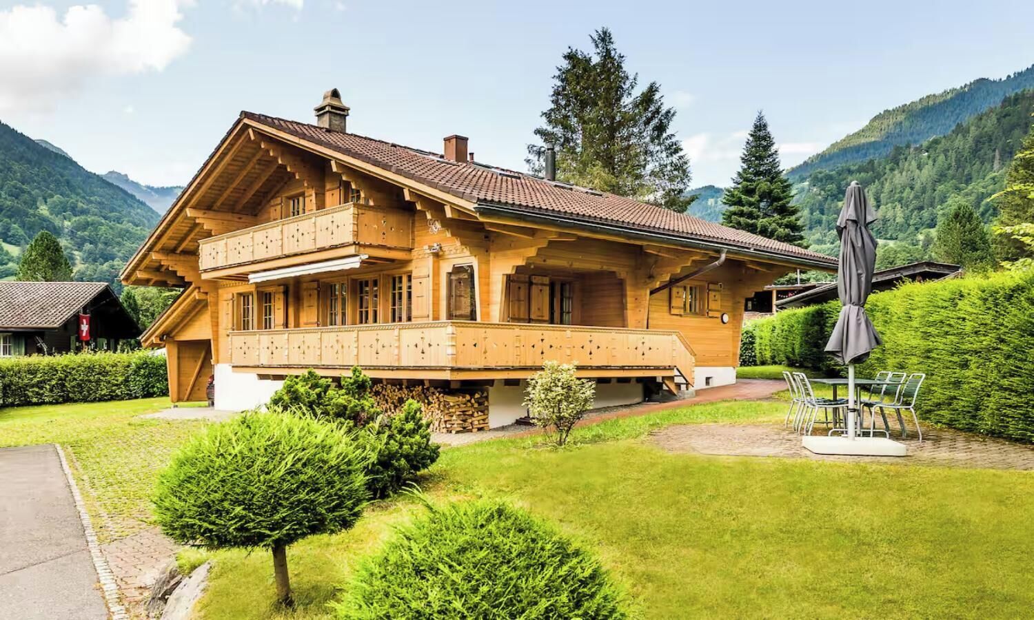 A chalet-style building with a balcony, surrounded by lush greenery and mountains.