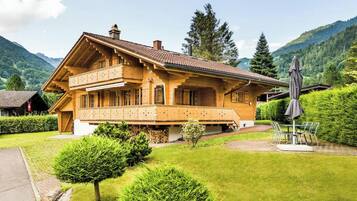 A chalet-style building with a balcony, surrounded by lush greenery and mountains.