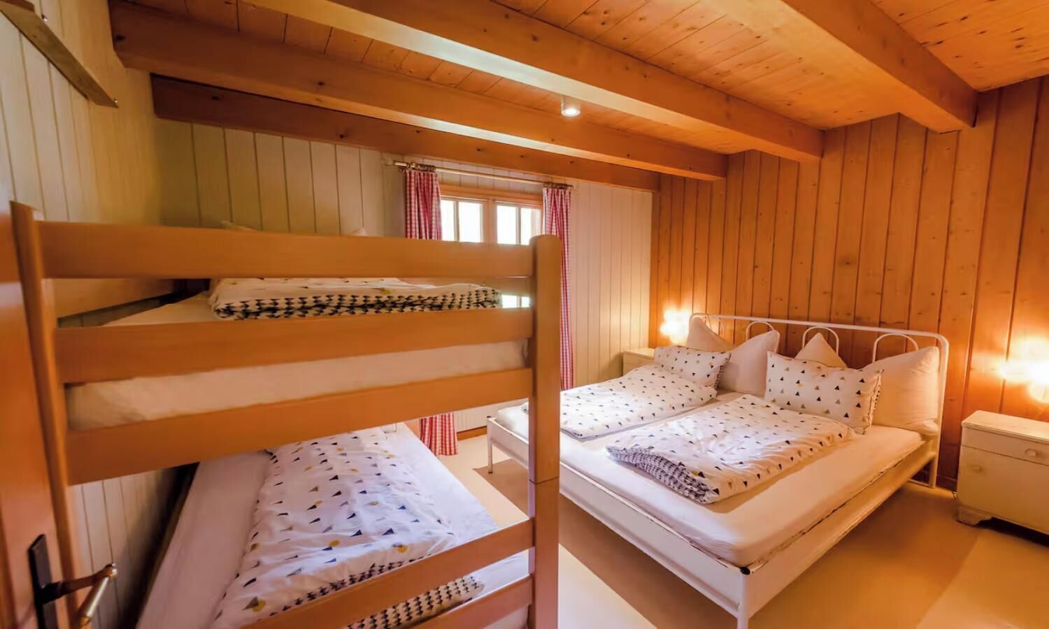 A wooden cabin bedroom with bunk beds and a single bed, a nightstand, and a window with curtains.