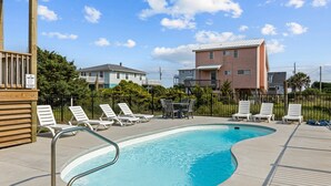 A swimming pool with lounge chairs and a table, surrounded by a fence and buildings in the background. - Waverlie’s Watch | Private Pool + Ocean and Sound Views (Emerald Isle)