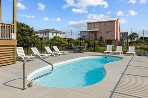 A swimming pool with lounge chairs and a table, surrounded by a fence and buildings in the background. - Waverlie’s Watch | Private Pool + Ocean and Sound Views (Emerald Isle)