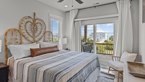 A bedroom with a bed, a ceiling fan, a window with curtains, and a view of a building. - Waverlie’s Watch | Private Pool + Ocean and Sound Views (Emerald Isle)