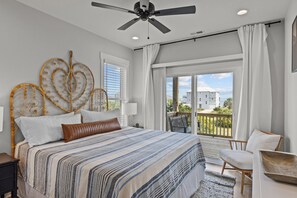 A bedroom with a bed, a ceiling fan, a window with curtains, and a view of a building. - Waverlie’s Watch | Private Pool + Ocean and Sound Views (Emerald Isle)