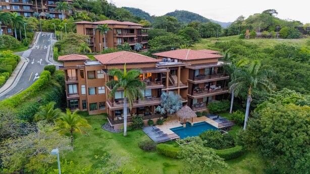 Un hotel de varios pisos con piscina, rodeado de abundante vegetación y una carretera.