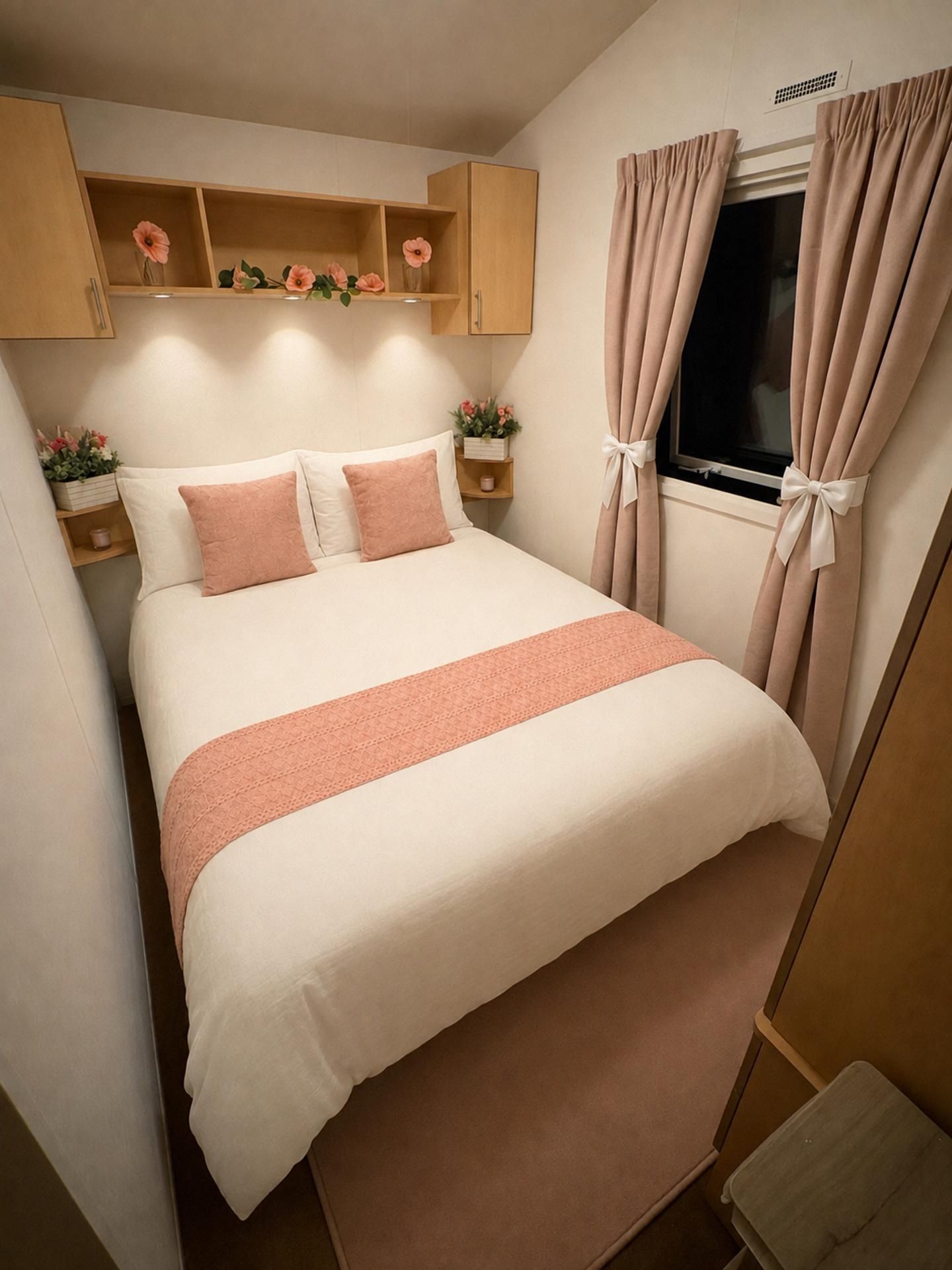Un dormitorio ordenado con cama, almohadas, una mesita de noche, una ventana con cortinas y una estantería con flores.