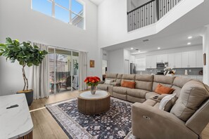 A spacious living area with a large sofa, a coffee table with flowers, and a view of the kitchen. - 5BR Pool & Spa Home with Themed Rooms 376 (KISSIMMEE)