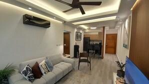 Ein modernes Wohnzimmer mit einer Couch, einem Deckenventilator, eingelassener Beleuchtung und einer Küche im Hintergrund.