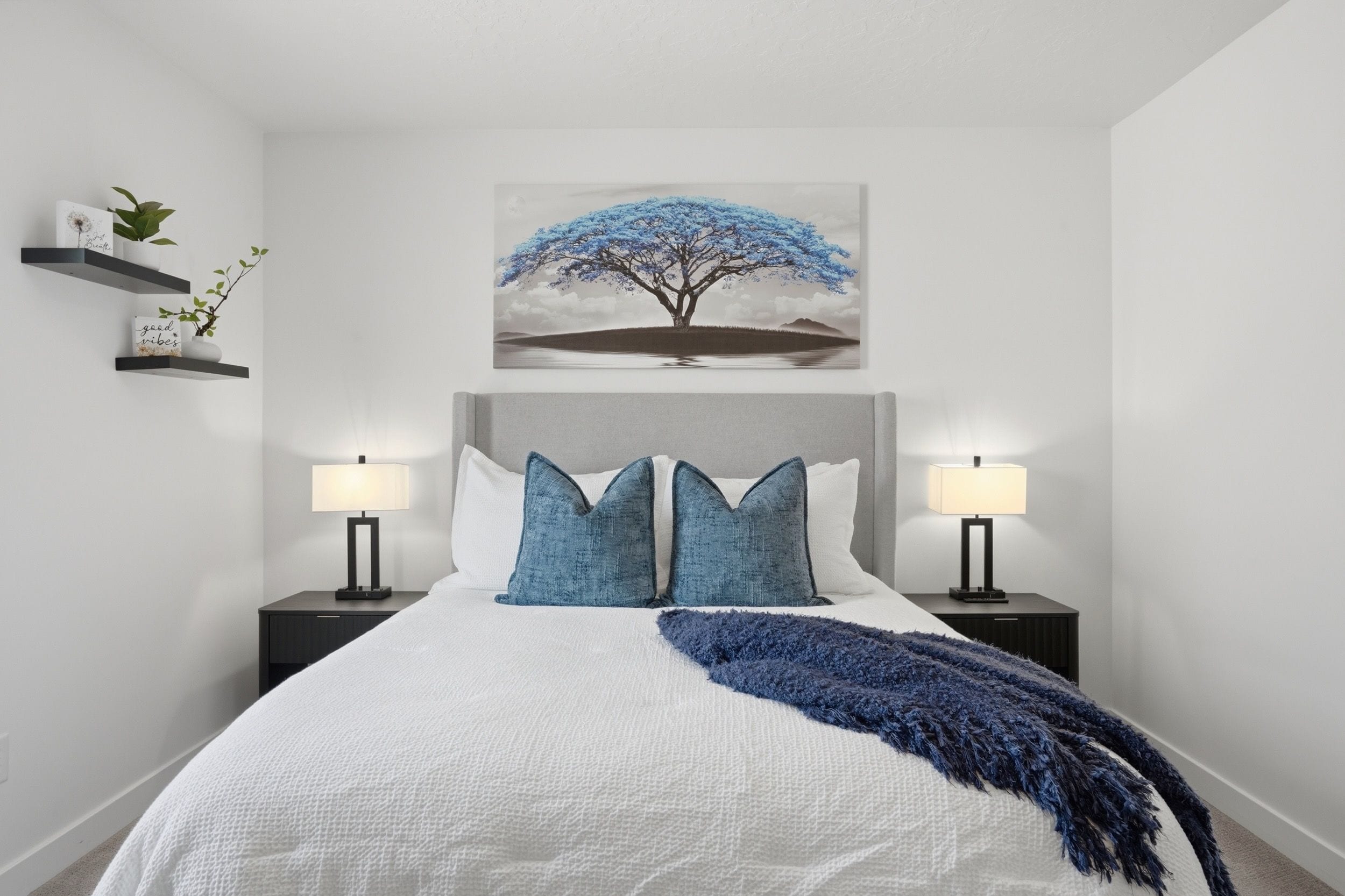 A bedroom with a large bed, two bedside tables with lamps, a wall-mounted shelf with plants, and a painting of a tree.