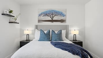 A bedroom with a large bed, two bedside tables with lamps, a wall-mounted shelf with plants, and a painting of a tree.