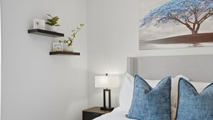 Une chambre à coucher avec un lit, des oreillers, une table de chevet, une lampe, une étagère murale ornée de plantes et un tableau.