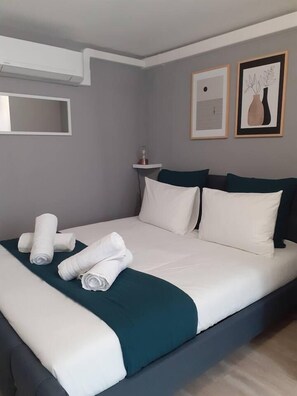 Un lit bien fait, avec du linge de lit blanc et un plaid turquoise, une petite étagère avec une bouteille, et deux œuvres d’art encadrées accrochées au mur.