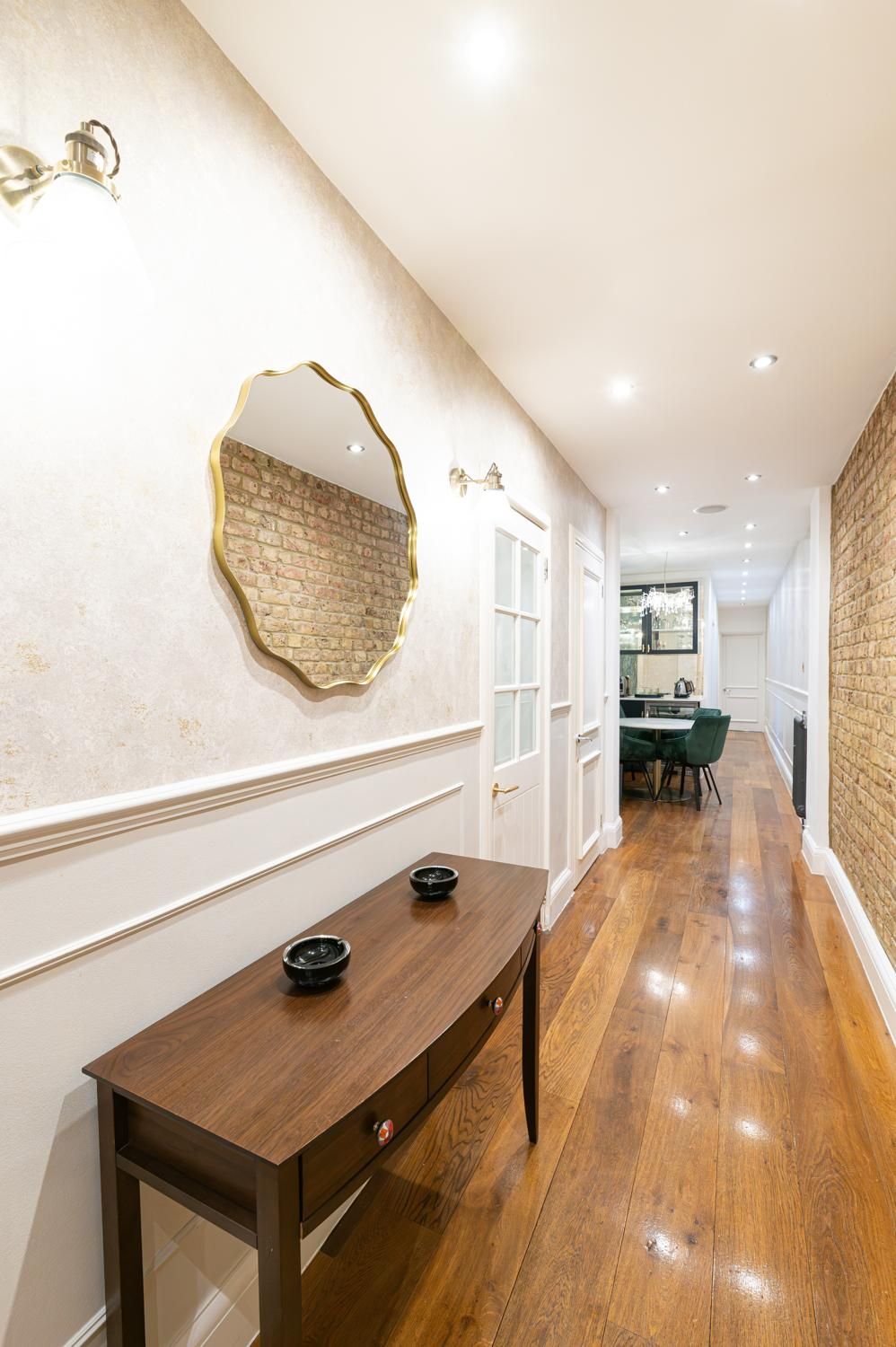 A hallway with a wooden floor, a dining table with chairs, a wall-mounted mirror, and a brick wall.