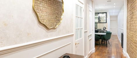 A hallway with a wooden floor, a dining table with chairs, a wall-mounted mirror, and a brick wall.