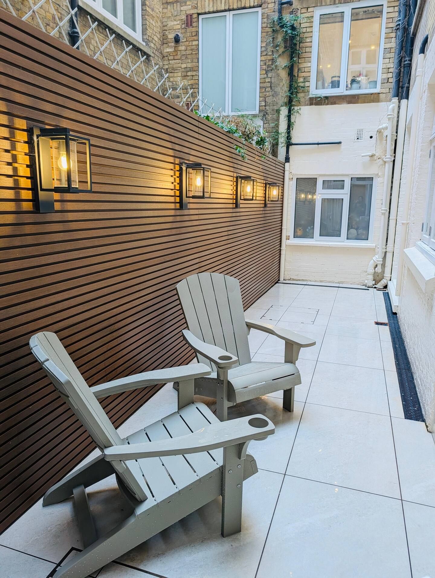 Un espace patio avec un banc en bois et deux fauteuils Adirondack, un sol carrelé et un mur en briques agrémenté d’une fenêtre.