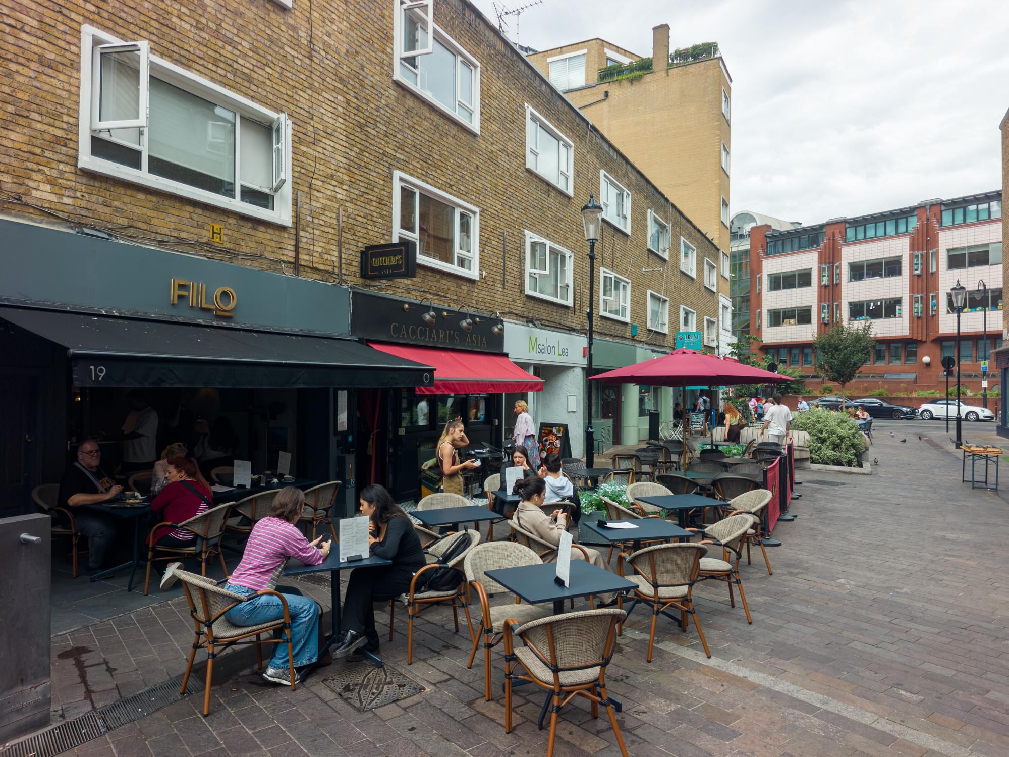 Un café en plein air avec des tables et des chaises, un bâtiment en briques abritant des boutiques, et un ciel dégagé.