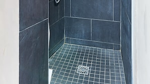 Zona de ducha con azulejos oscuros, cabezal de ducha de mano y grifo empotrado en la pared.