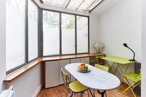 A compact kitchen with a large window, a dining table with chairs, and a small green table with a lamp. - Cozy Studio in the Heart of Montorgueil (Paris)