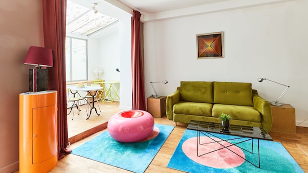 Una sala de estar con un sofá verde, un reposapiés rosa, una mesa de centro de vidrio y una lámpara roja.