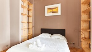Una cama bien tendida con sábanas blancas y una toalla enrollada, un cabecero negro y estanterías de madera a ambos lados.
