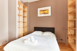 Un lit bien fait, avec du linge de lit blanc et une serviette enroulée, une tête de lit noire et des étagères en bois de chaque côté.