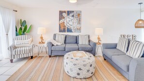 A living room with a grey sofa, striped armchairs, a round ottoman, and a large abstract painting on the wall. - A-Wave for Everyone - Full duplex + 2 Pools! (New Smyrna Beach)
