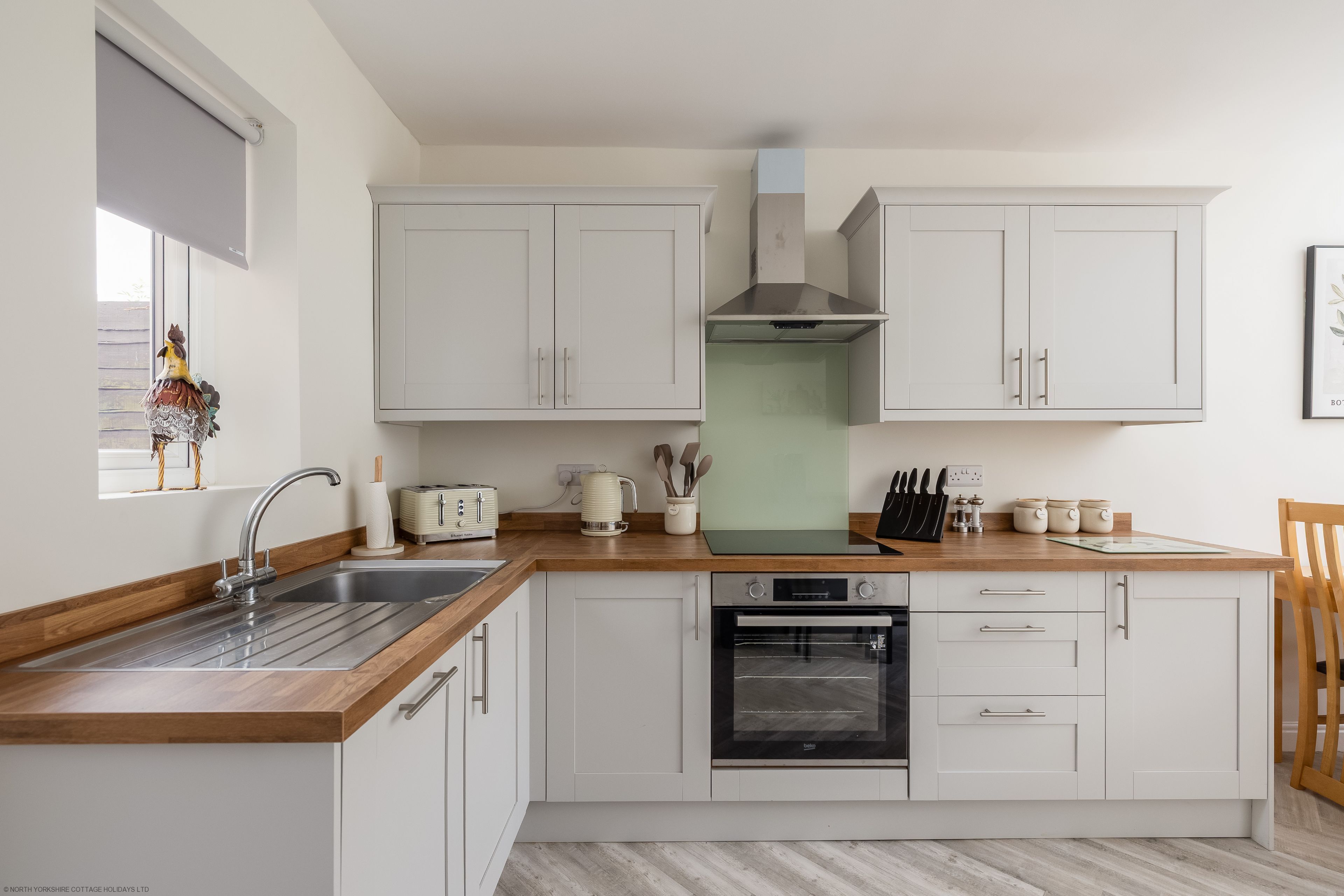 Una cucina moderna con mobili bianchi, un piano di lavoro in legno, un forno integrato e una finestra con un gallo decorativo.
