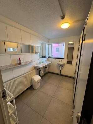 Une petite salle de bain comprenant des toilettes, un lavabo et un miroir. Une fenêtre laisse entrer la lumière naturelle et un radiateur est fixé au mur.