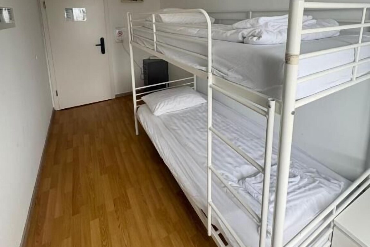 Ein Schlafraum mit Etagenbetten, Holzboden, weißen Wänden und einer Tür im Hintergrund.
