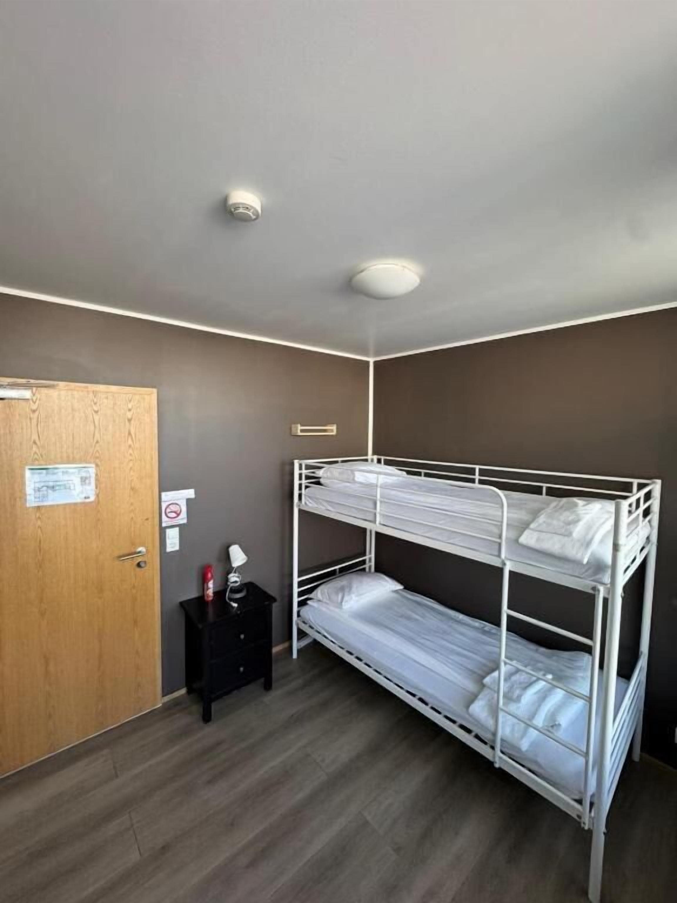 Una habitación con literas, una puerta de madera, una mesita con una lámpara e iluminación en el techo.