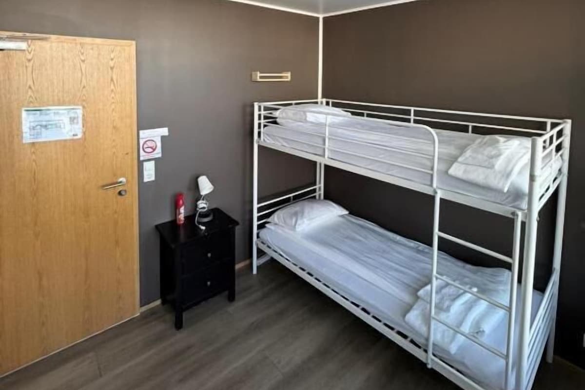 Ein Zimmer mit einem Etagenbett, einer Holztür, einem kleinen Tisch mit Lampe und einer Deckenleuchte.