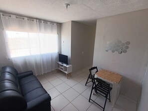 A living room with a black sofa, a small TV on a stand, a dining table with chairs, and a window with sheer curtains. - Apartment with two bedrooms in the bedroom (Macapá)
