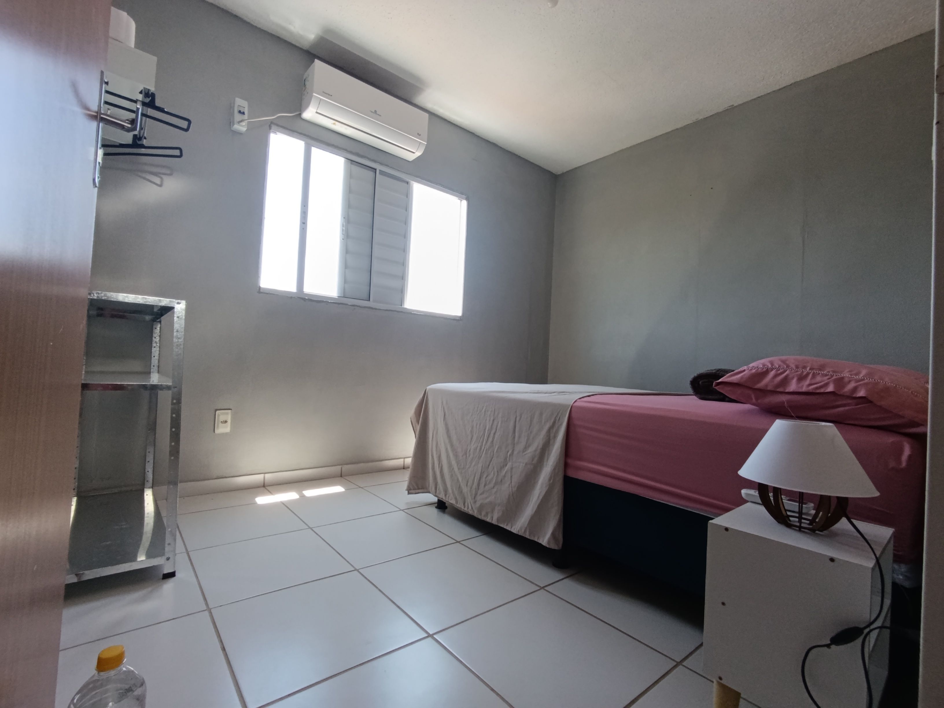 A bedroom with a bed, a window, an air conditioner, and a bedside lamp.
