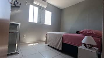 A bedroom with a bed, a window, an air conditioner, and a bedside lamp.