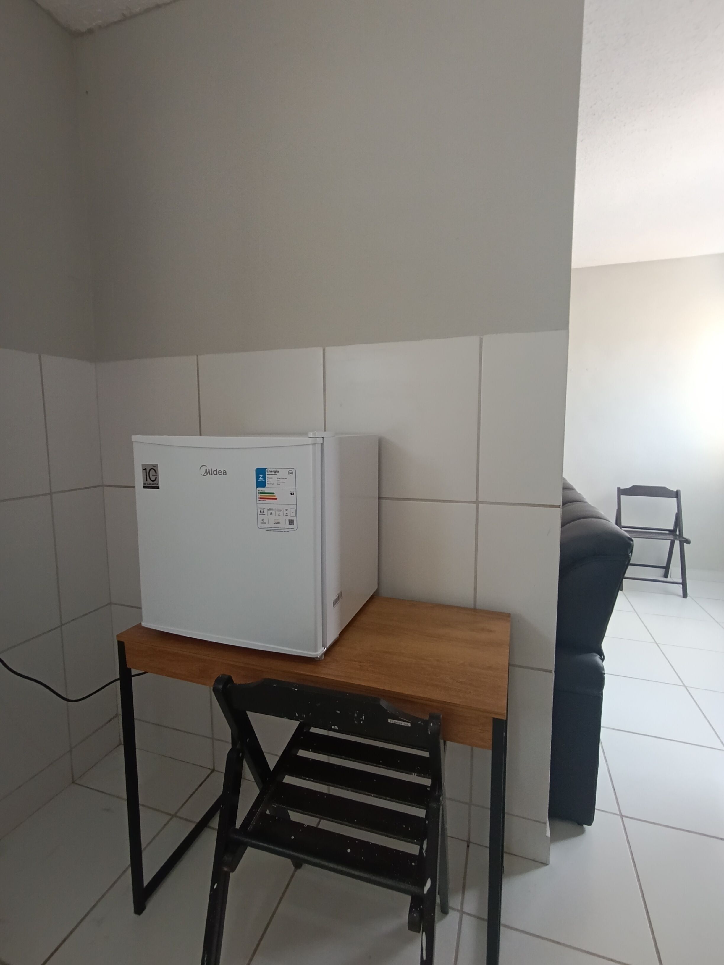 Quarto pequeno com geladeira branca, mesa de madeira, cadeira preta e sofá cinza.