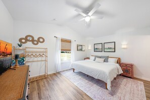 Ein Schlafzimmer mit Bett, Deckenventilator, Fenster mit Vorhängen, Nachttisch und Wandbild.