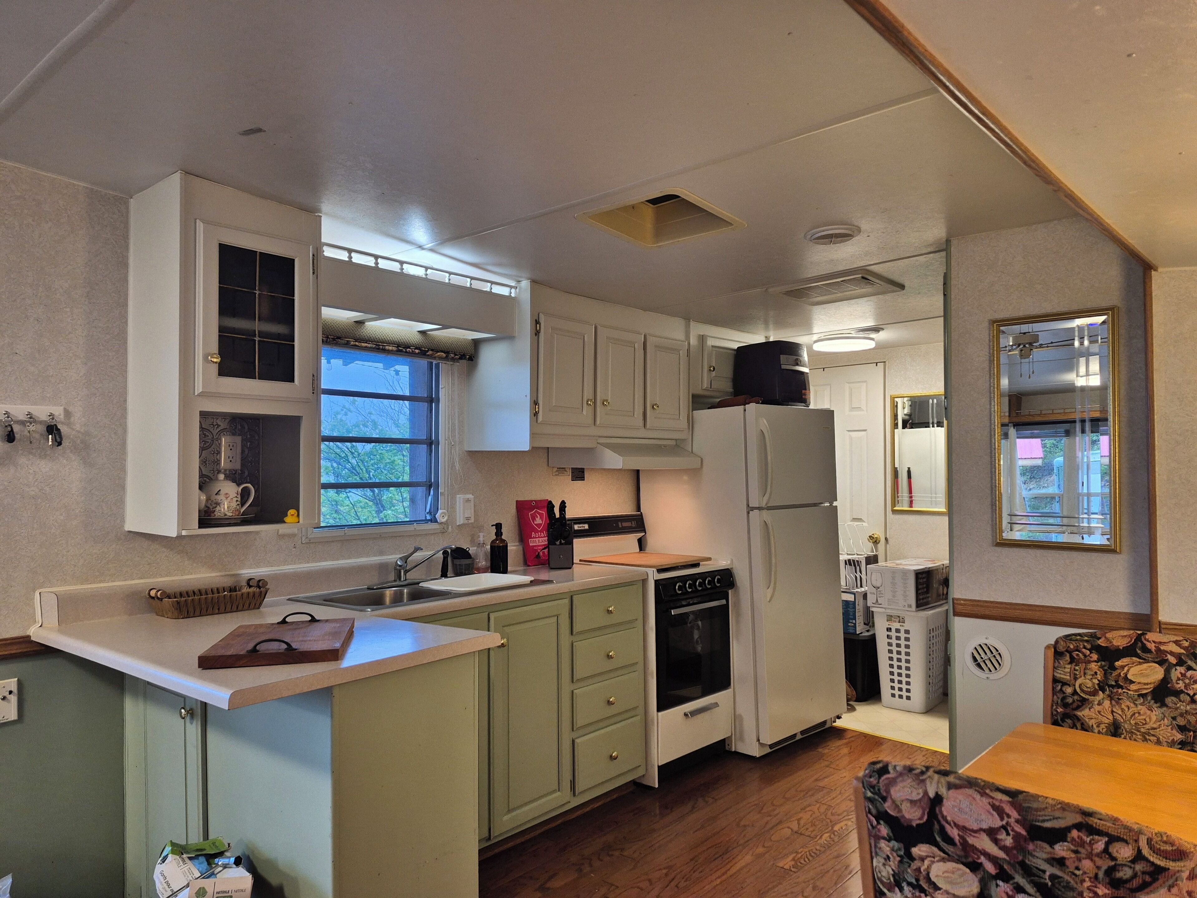 A compact kitchen with a refrigerator, oven, and sink. There is a window providing natural light and a small dining area with a floral-patterned chair.